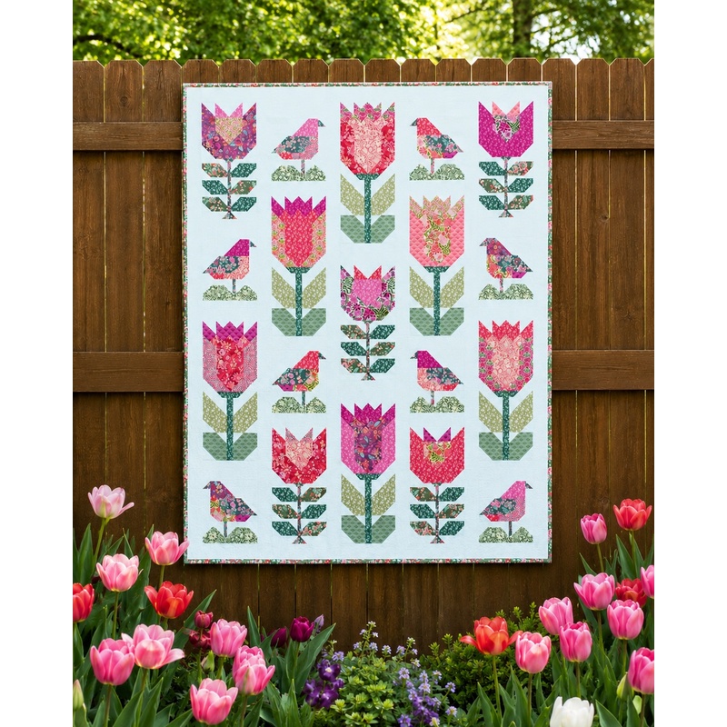 The Birds and Blooms quilt, colored in bright fabrics from the Floriori collection by Moda Fabrics and staged on a wooden fence in a garden of tulips.