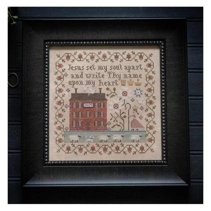 The completed cross stitch project featuring a brick house, a shepherd leading sheep, a bible phrase, and a floral border, in a black frame.