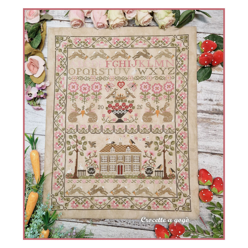 Finished April Sampler cross stitch pattern on a beige fabric surrounded by berries, florals, and carrots.