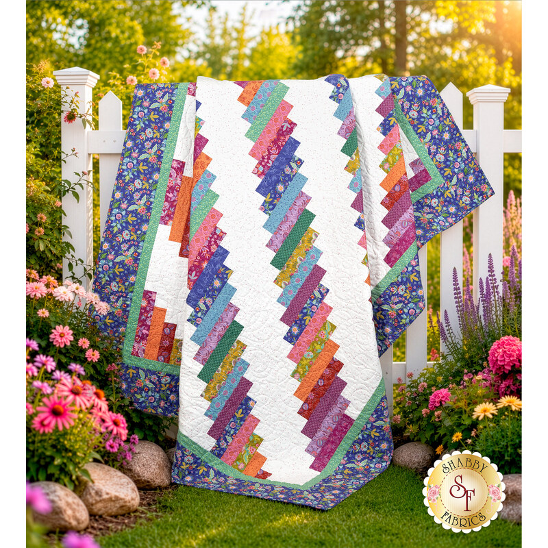 The completed 28th Floor Quilt colored in white blue, and a rainbow of fabrics from the Kipling Gardens collection by Riley Blake Designs, artfully draped over a white fence in a lush garden.