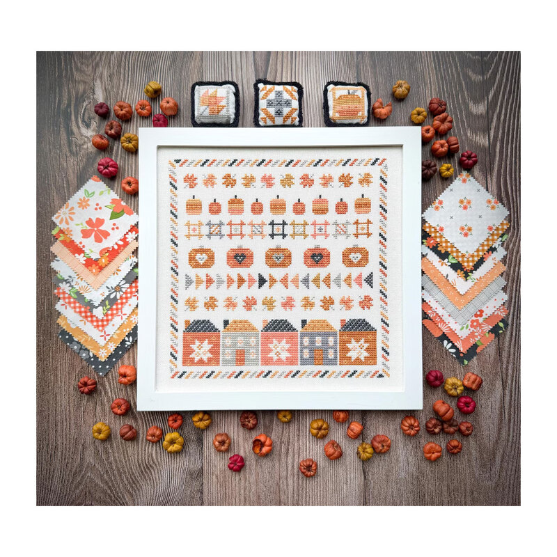 Finished Pumpkin Hollow cross stitch pattern inside of a white frame surrounded by small pumpkins, fabrics from Moda's Birch and Willow fabric line, and smaller accent pillows.