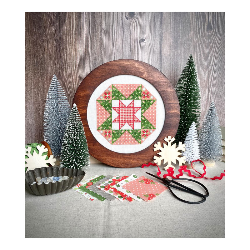 The Whimsy Star cross stitch pattern showing the finished product in a wooden circle hoop next to frosty prop trees, a snowflake, the Moda Poinsettia and Pear line, and a tub of buttons.