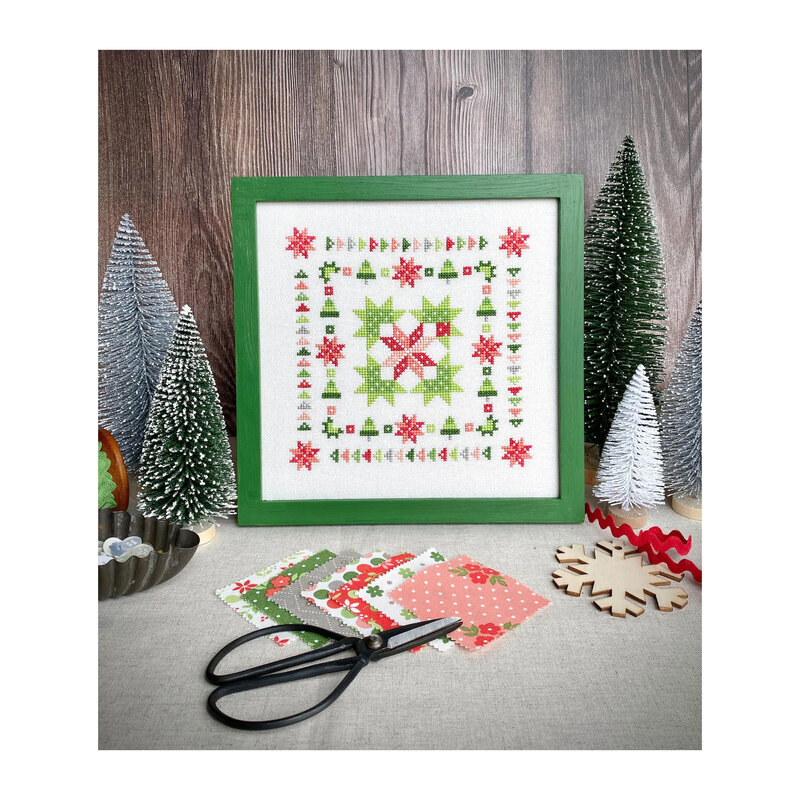 Finished Wreath and Pine cross stitch pattern in a green square frame surrounded by frosted pine trees, a snowflake, Moda's Poinsetta and Pear fabrics, and a tub of buttons.