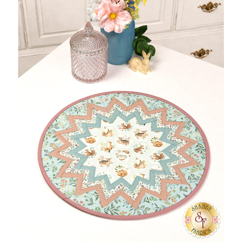 The completed Point of View Folded Star Table Topper, colored in serene shades of blue, pink, and cream fabrics from the Woodland Heart collection by Northcott Fabrics, staged on a white table with coordinating décor.
