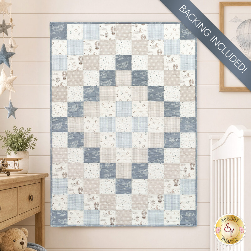 The completed Trip Around The World Crib Quilt colored in blue, taupe, and white fabrics from the Soaring Dreams collection by FIGO Fabrics, staged on a taupe wall in a nursery with a crib and set of drawers. A banner in the corner reads "Backing Included!"