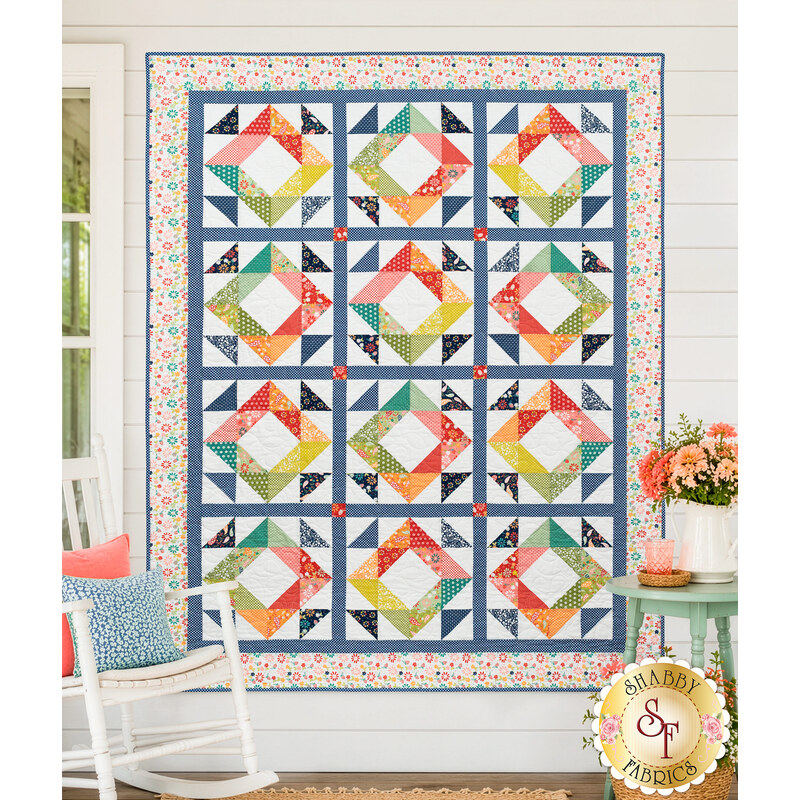 The completed Mosaic Diamonds Quilt, colored in a rainbow of prints from the Raspberry Summer collection by Moda Fabrics, staged on a white porch wall with coordinating décor and furniture.
