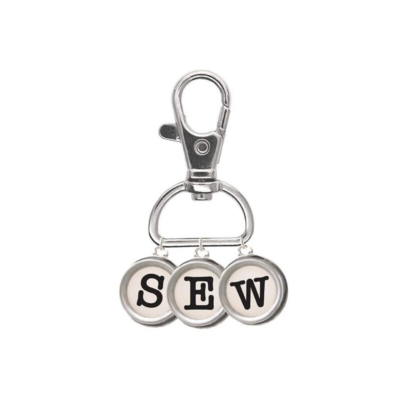 Enamel pin with a clip holding three small circular charms spelling out the word "SEW" in a typewriter typeface isolated on a white background.