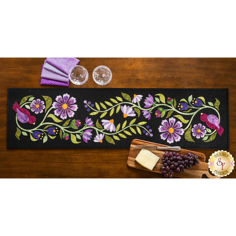 The completed Baltimore Garden Table Runner in violet, staged on a wooden table with four black chairs, two wine glasses, fabric, and a serving plate with cheese and grapes.