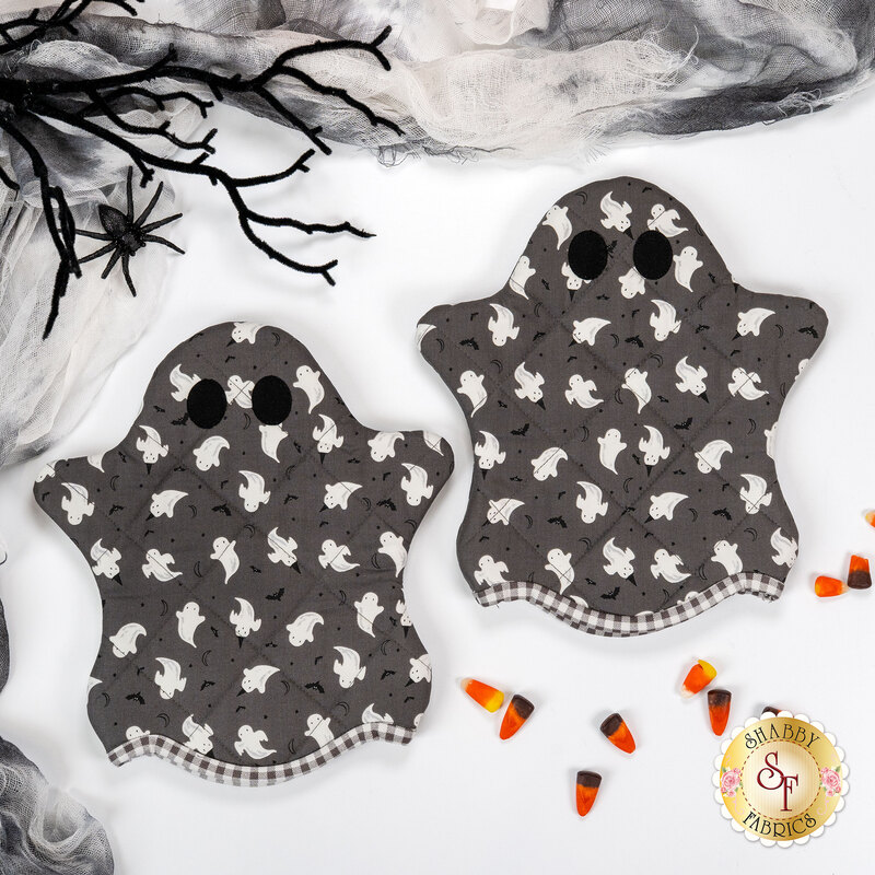 The two completed Ghostly Hot Pads, colored in gray and white glow in the dark fabrics from the Frank and Friends collection, staged on a white table with coordinating décor and candy corn.