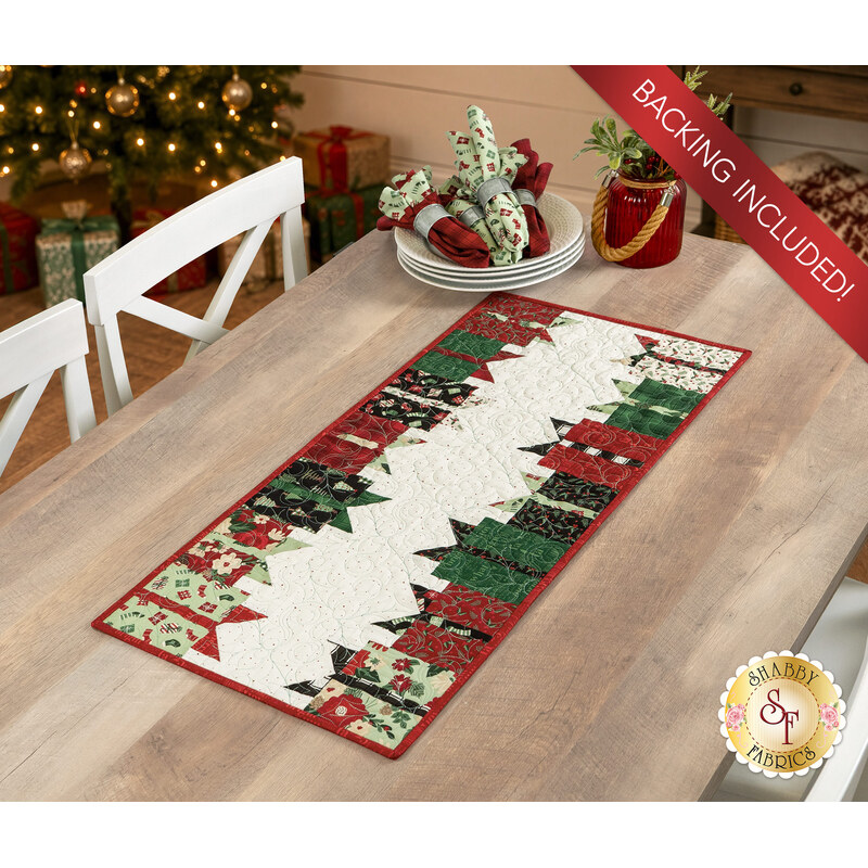 The completed A Gift for You runner colored in red, cream, and mint green fabrics from the Evergreen Wishes collection by Riley Blake Designs, staged on a wooden table with coordinating plates and flowers in a room decorated for Christmas. 