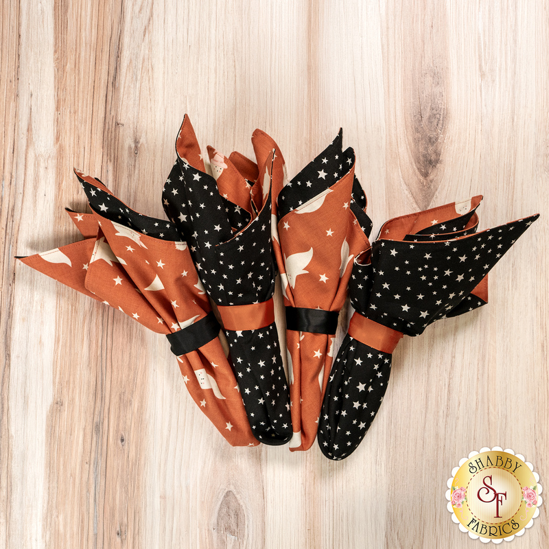 A top-down shot of the four napkins colored in the Purrs and Boos collection from Moda Fabrics, featuring cute Halloween themed prints, staged on a wooden table.