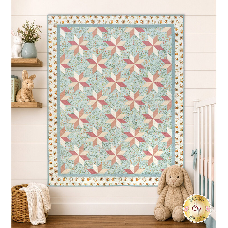 The completed Star of the Show Baby Quilt, colored in serene shades of blue, pink, and cream fabrics from the Woodland Heart collection by Northcott Fabrics, staged on a cream shiplap wall in a nursery with coordinating stuffed animals and décor.
