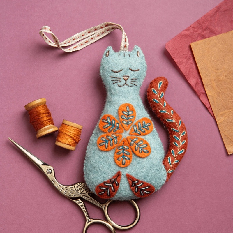 Handmade felt cat with embroidered flowers, scissors, thread spools, and paper on pink background.