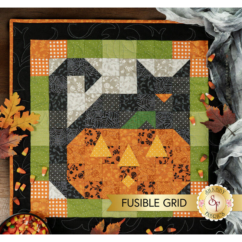 A top down shot of the first block of the Mystery BOM, a Fusible Grid patchwork block of a black cat with an orange pumpkin with yellow laser cut appliqué pieces, staged with coordinating leaves, spooky cobwebs, and candy corn. A gold banner with the Shabby Fabrics logo reads "Fusible Grid."
