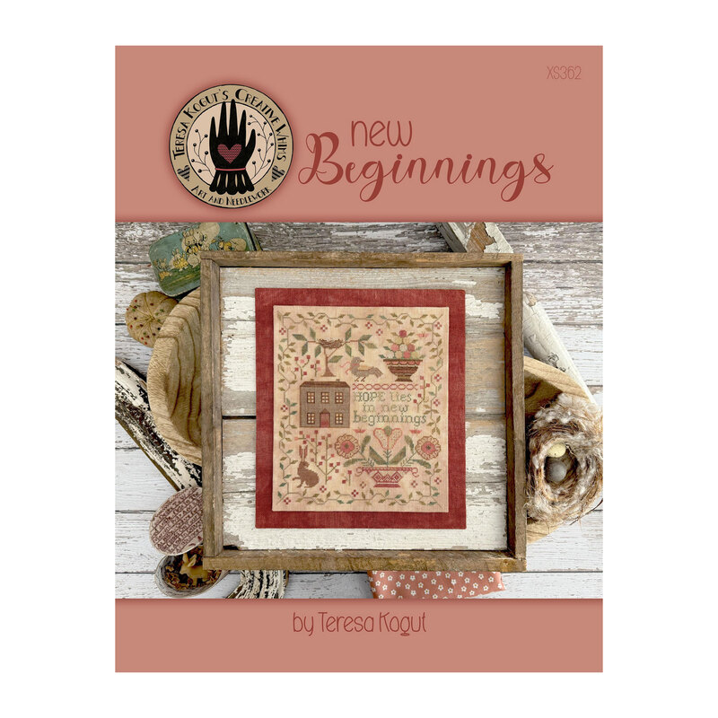Front page of the New Beginnings cross stitch pattern finished in a wooden frame next to quaint house supplies and a birds nest.