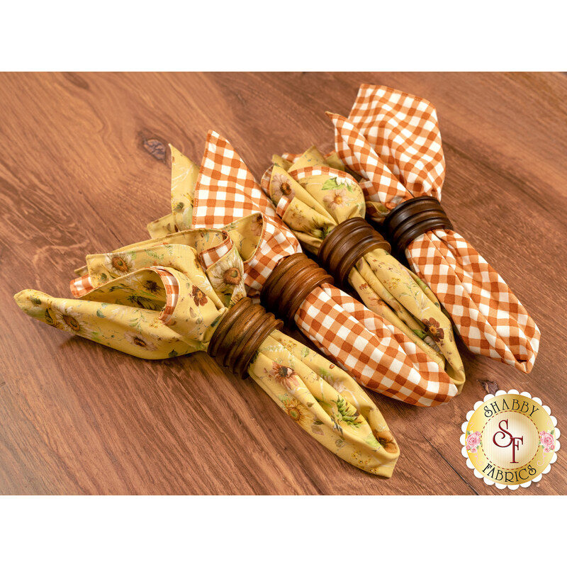 The four completed cloth napkins, colored in yellow and orange fabrics from the Sunflower Whispers collection by Riley Blake Designs, staged on a warm wooden table.