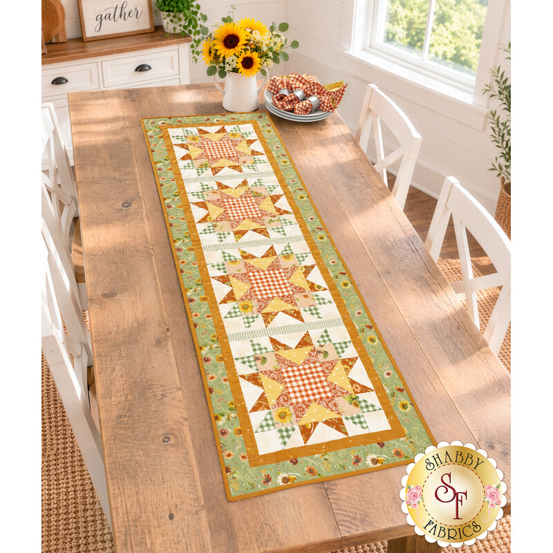 The completed Fancy Star Table Runner, colored in warm fabrics from the Sunflower Whispers collection by Riley Blake Designs, staged on a wood table with coordinating décor, white chairs, speckled with sunlight.