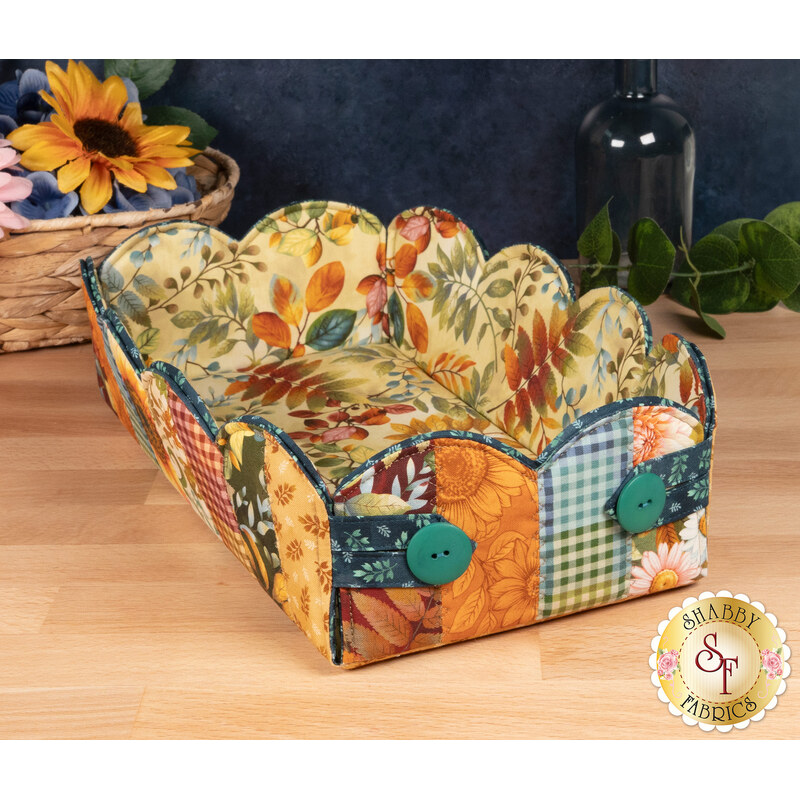 A shot of the completed Scalloped Basket, colored in warm autumnal fabrics from the Baskets of Autumn Blessings from Studio E Fabrics, staged on a wooden table with coordinating décor.