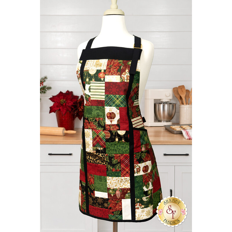 The completed Charm Squares Apron, colored in red, green, cream, and gold metallic fabrics from the Poinsettias at Christmas collection by Jennifer Bosworth for Riley Blake Designs, staged on a mannequin in an airy kitchen space.