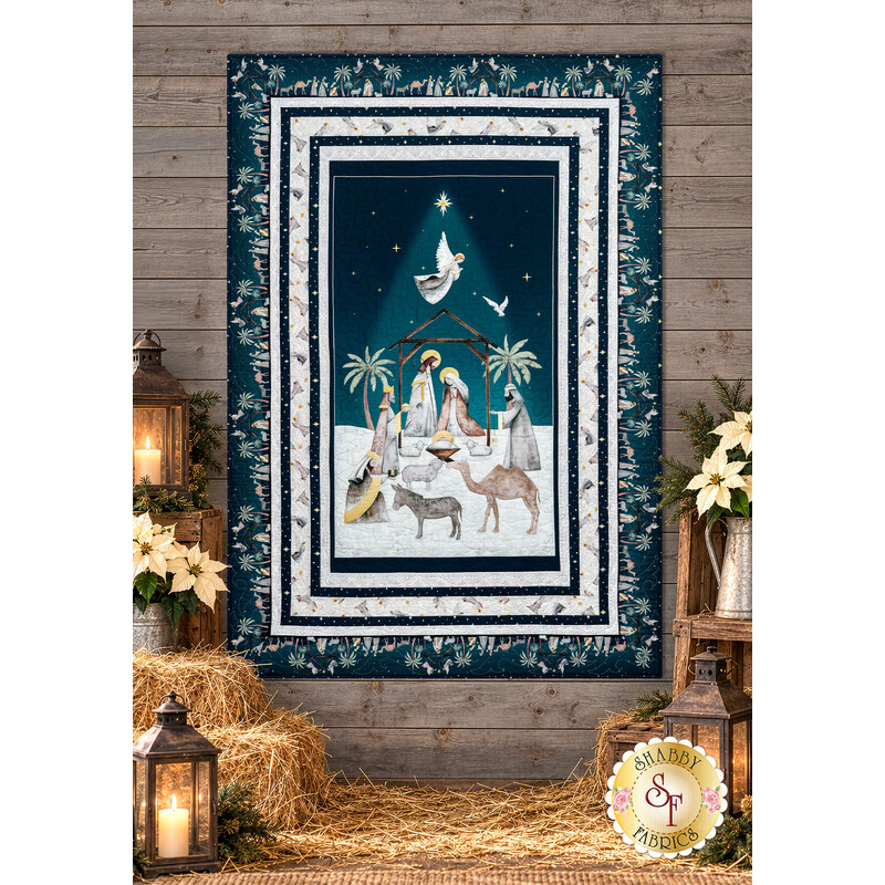 The completed Divine Reunion Quilt, colored in navy blue and gray/white fabrics from the Silent Night collection by Blank Quilting, hung on a rustic wood wall and staged with hay, lanterns, and white poinsettias.