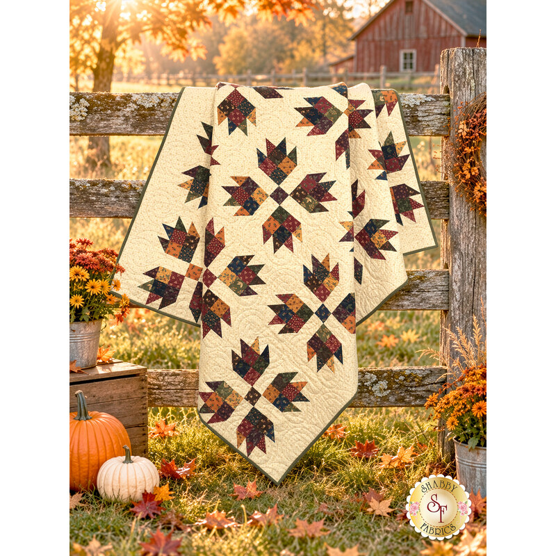 The completed Bear Hugs quilt, colored in warm flannels from the Flannel Season collection by Moda Fabrics, artfully draped over a rustic wood farm fence with a barn in the background and pumpkins and florals in the foreground.