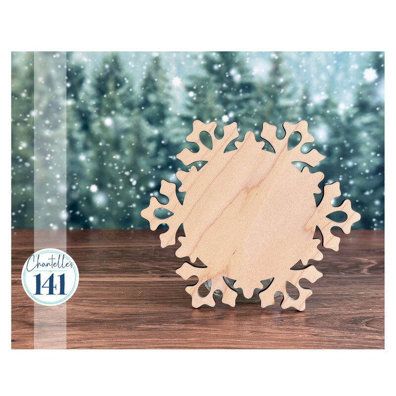 The unfinished snowflake ornament, staged with a snow-frosted pine forest background on a wood surface.