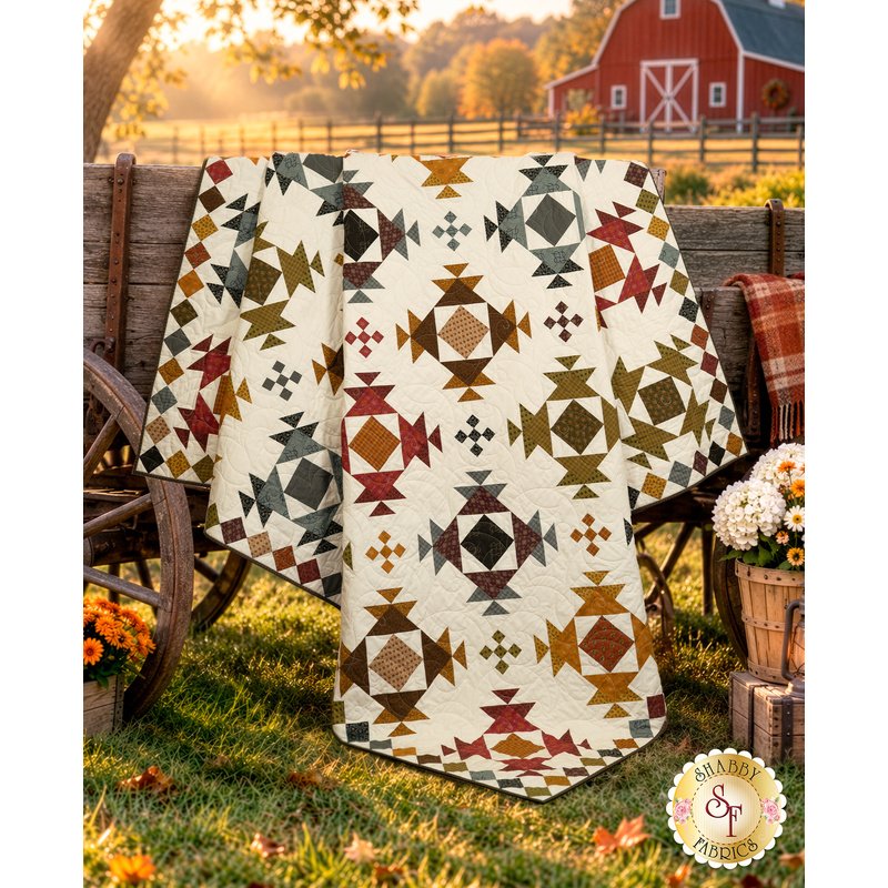 The completed Harrisonburg Quilt,  colored in rich fabrics from the Jamestown collection from Andover Fabrics, artfully draped over a rustic wooden wagon and coordinating flowers; a red barn can be seen in the background.