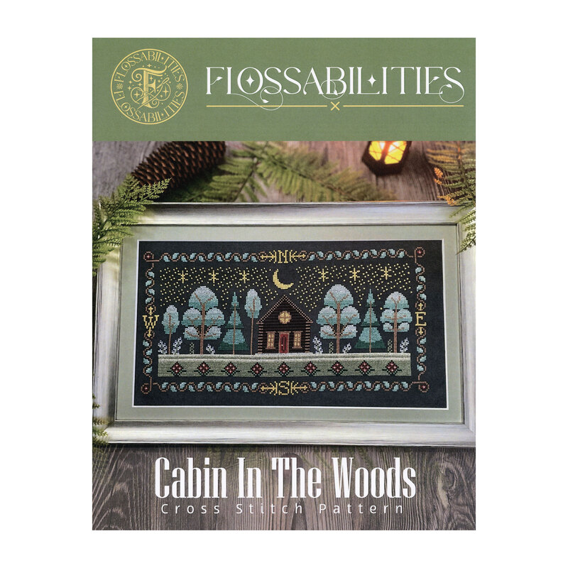 Front cover of the Cabin in the Woods cross stitch booklet with the finished pattern in a frame on a wooden table.