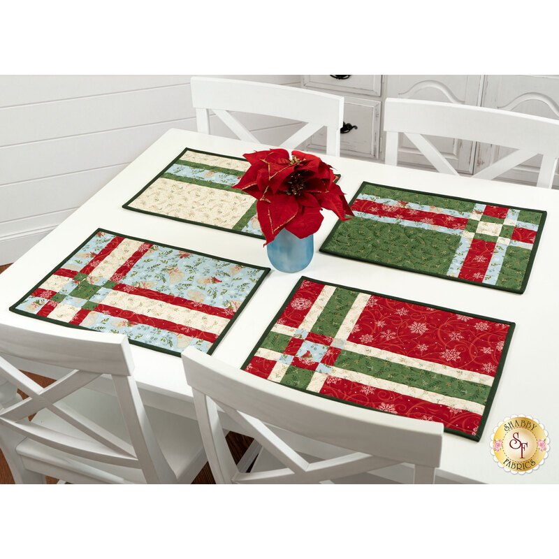The completed Fat Quarter Shuffle Mats, colored in red, green, navy, light blue, cream, and white fabrics from the When Snow Falls collection from Moda Fabrics, staged on a white table with four settings and a blue jar of poinsettias in the center.