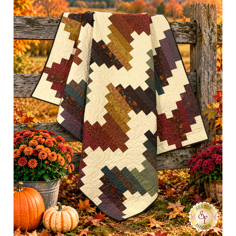 The completed Ridiculously Easy Jelly Roll Quilt, colored in warm, rustic prints from the Bountiful Blessings collection by Henry Glass Fabrics and artfully draped over a wooden fence, staged with rich coordinating florals and pumpkins.