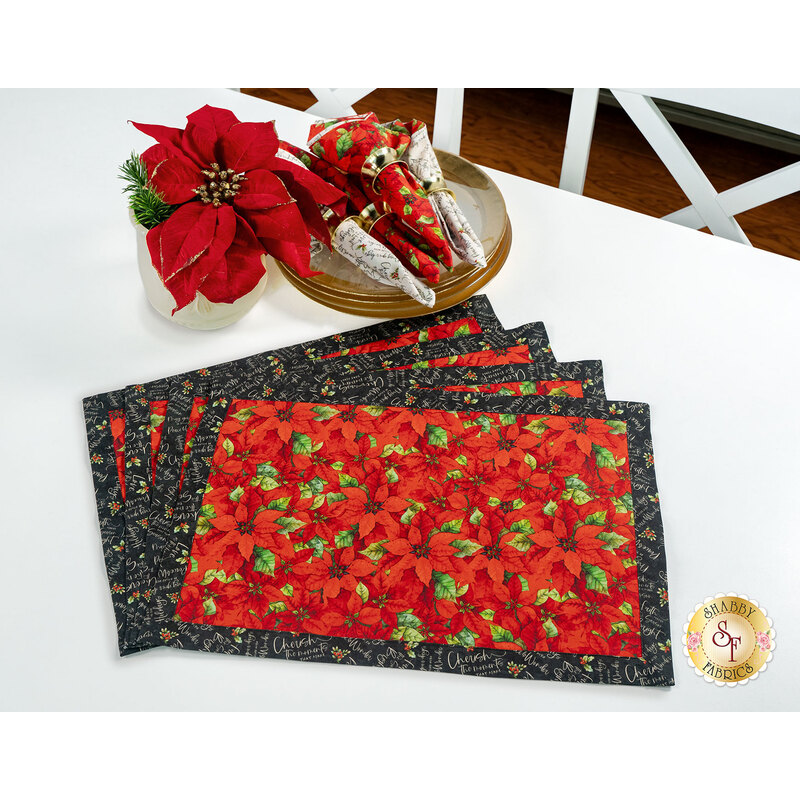 The four completed Self-Binding Placemats, fanned out and  colored in red, cream, and black fabrics from the Poinsettia Radiance collection from White Owl Textiles, staged on a white table with coordinating décor.
