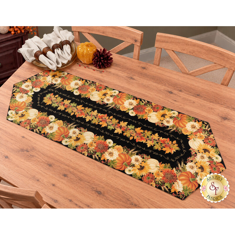 The completed Easy Striped Table Runner, colored in warm, autumn themed prints from the Give Thanks collection by Timeless Treasures Fabrics, staged on a warm wooden table with coordinating décor and table settings.