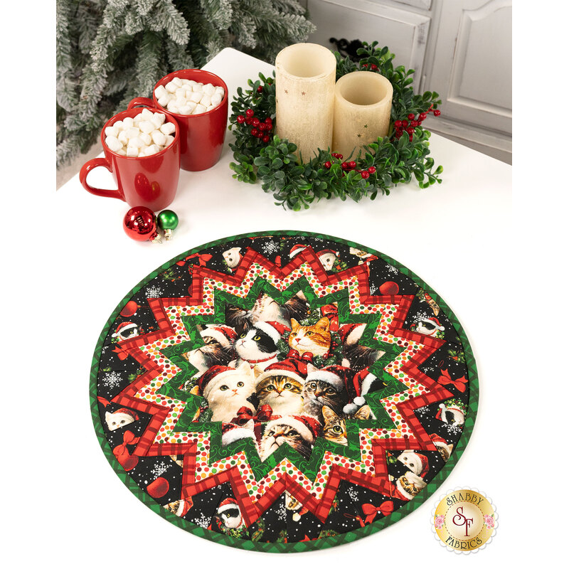 The completed Point of View Folded Star Table Topper colored in black, red, and green fabrics from the Merry Cats collection by Benartex, staged on a white table with coordinating décor and cups of cocoa.