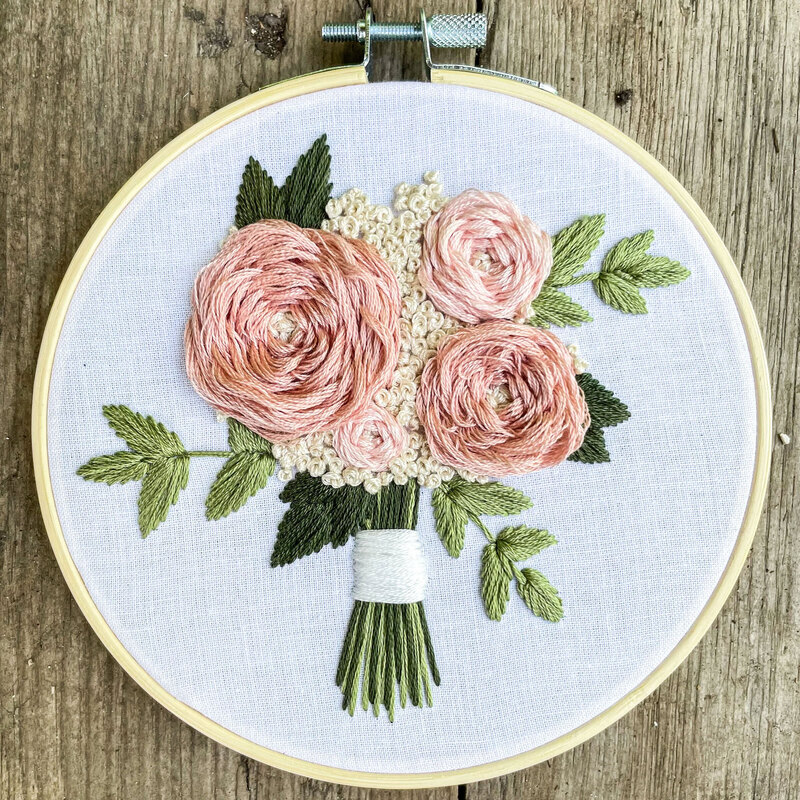 Round embroidery hoop displaying a hand-stitched bouquet of pink roses and green leaves on light fabric.