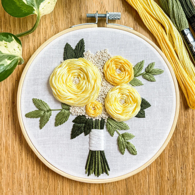Round embroidery hoop displaying a hand-stitched bouquet of yellow roses and green leaves on light fabric.