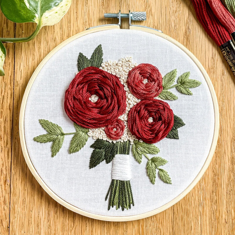 Round embroidery hoop displaying a hand-stitched bouquet of red roses and green leaves on light fabric.