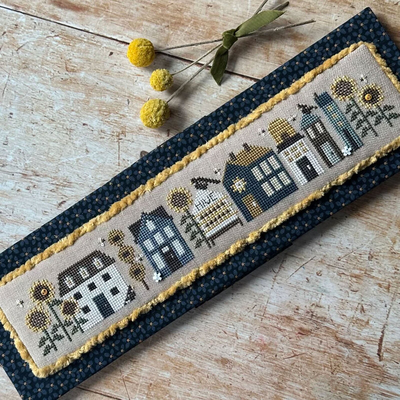 A cross stitch village with whimsical houses surrounded by bees, beehives, sunflowers, and honey-inspired details.