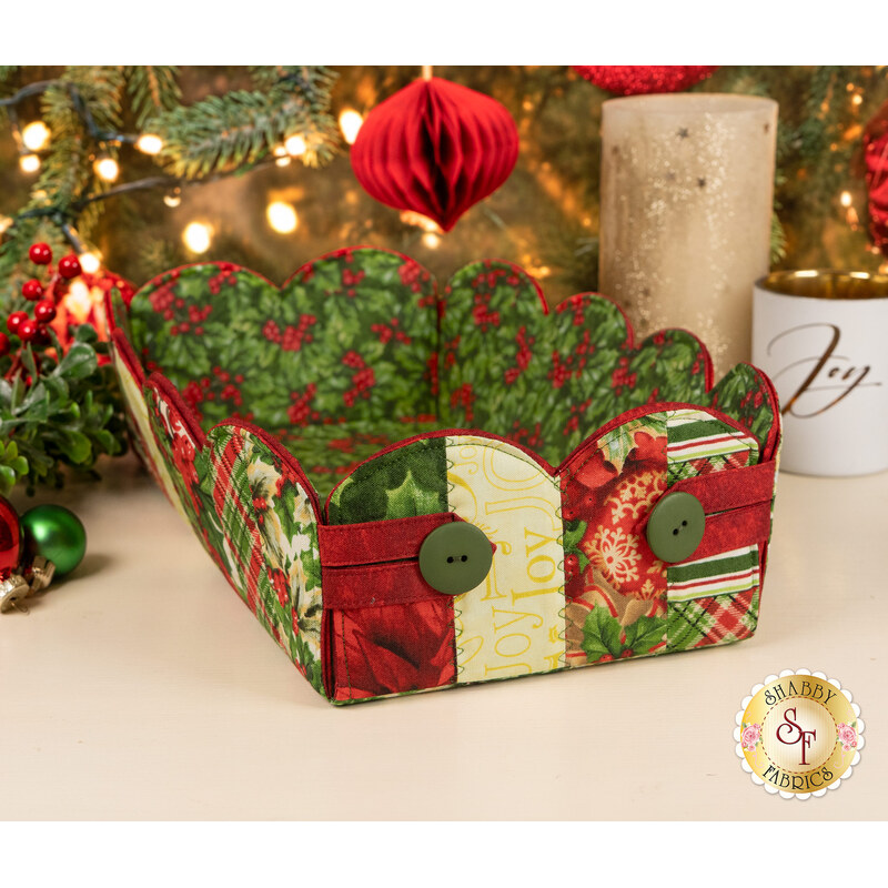 The completed Scalloped Basket, colored in cream, red, and green fabrics from the Deck the Halls collection by Northcott Fabrics, staged on a cream table with coordinating flowers and décor.