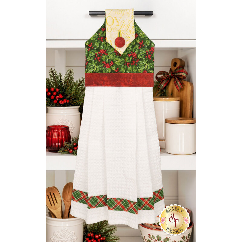 The completed Hanging Towel colored in cream, red, and green fabrics from the Deck the Halls collection by Northcott Fabrics, staged on a kitchen drawer with coordinating decor.
