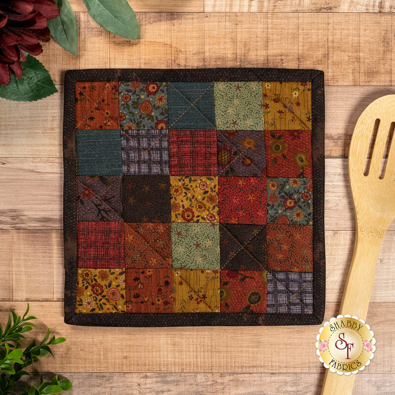 A top down shot of the completed Fusible Grid Hot Pad, colored in warm, rustic prints from the Bountiful Blessings collection by Henry Glass Fabrics, staged on a wooden countertop with coordinating flowers and a wooden spoon.