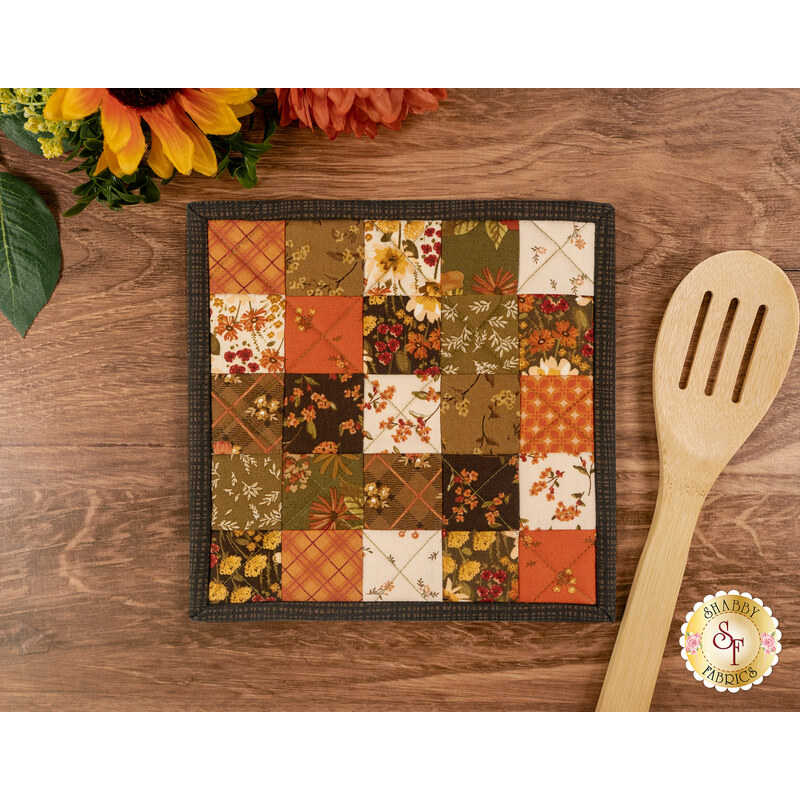 The completed Fusible Grid Hot Pad colored in autumn fabrics from the Bountiful collection by Riley Blake Designs, staged on a wooden surface with a wooden spoon.
