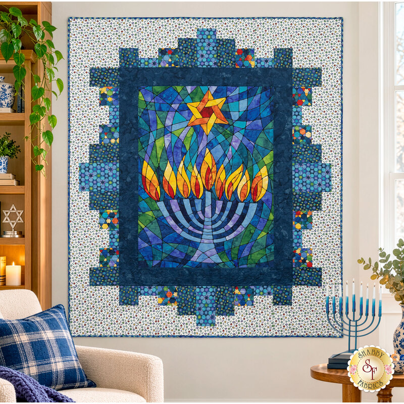 The completed Hanukkah Lights Quilt, colored in blue, yellow, green, and orange fabrics from the Hanukkah Lights collection by Northcott Fabrics, hung on a white wall and staged with coordinating décor and a festive hanukkiah/menorah.