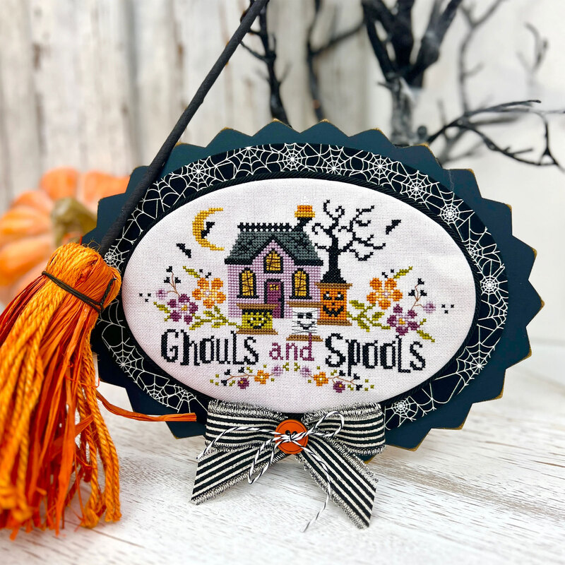 Black painted wooden cross stitch display with a mounted Halloween stitched piece, coordinating fabric, black trim, striped ribbon, twine, and a decorative button.