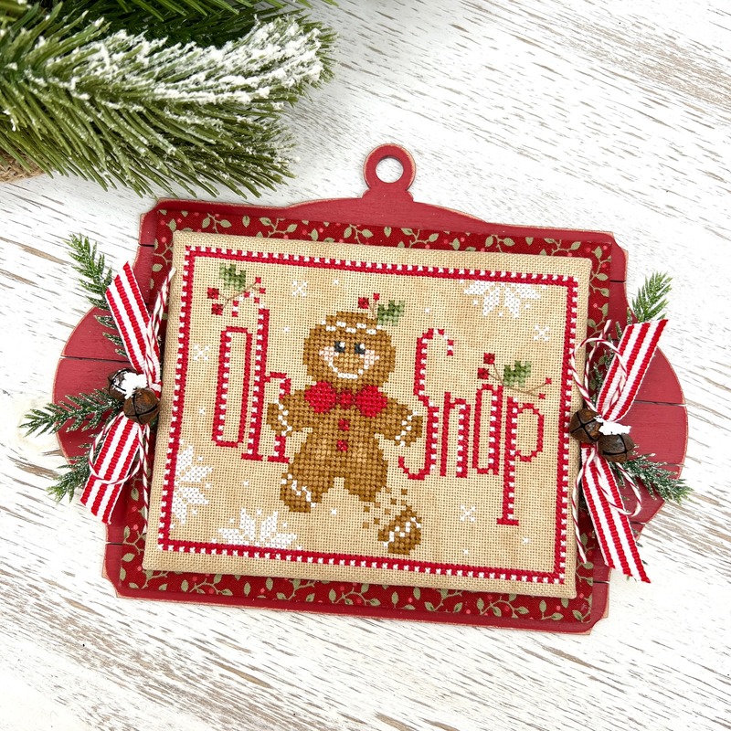 A Christmas cross stitch ornament with a gingerbread man, red bow, and candy cane-inspired lettering.
