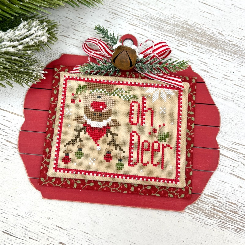 A Christmas cross stitch ornament with a wide-eyed reindeer wearing a green scarf, tiny ornaments hanging from its antlers, and a sprig of holly.