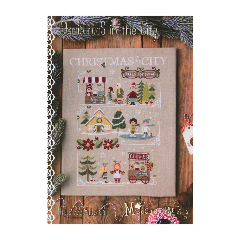 Front cover of the Christmas in the City cross stitch pattern showing the finished pattern laid on a wooden plank table on top of pine boughs, string, and Christmas gift tags.
