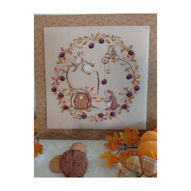 Photo of the finished Snack in the Woods cross stitch pattern placed on a floral fabric board next to pumpkins, cookies, and maple leaves.
