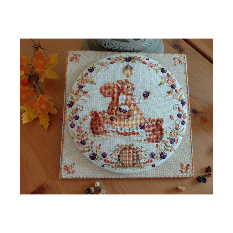 Photo of the finished Autumn Day - Squirrel cross stitch pattern laid on a wood plank table, lantern, and leaves.