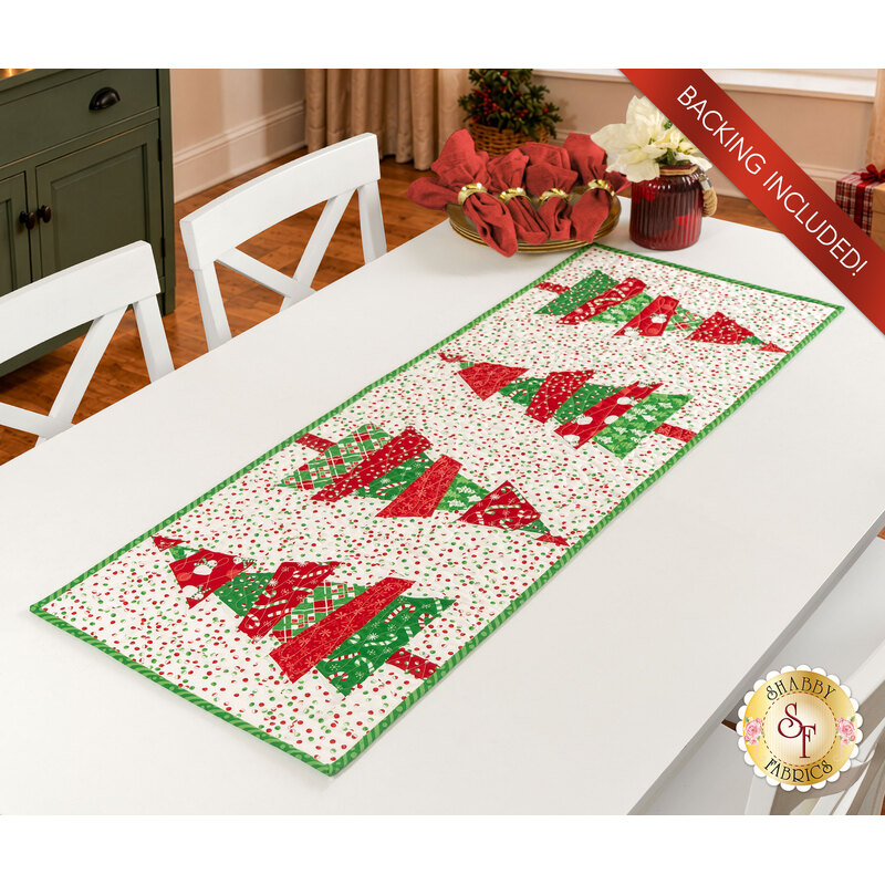 The completed Crazy Christmas Tree table runner, colored in bright red, green, and white fabrics from the Snow Time collection by Moda Fabrics, staged on a white table with coordinating napkins and décor.