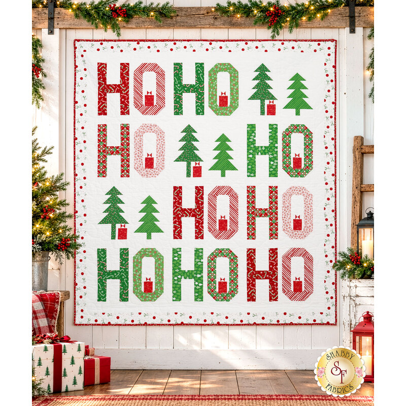 The completed Jollity quilt kit, colored in bright red, green, and white fabrics from the Snow Time collection by Moda Fabrics, staged on a white paneled wall and surrounded by coordinating garlands, presents, and décor.
